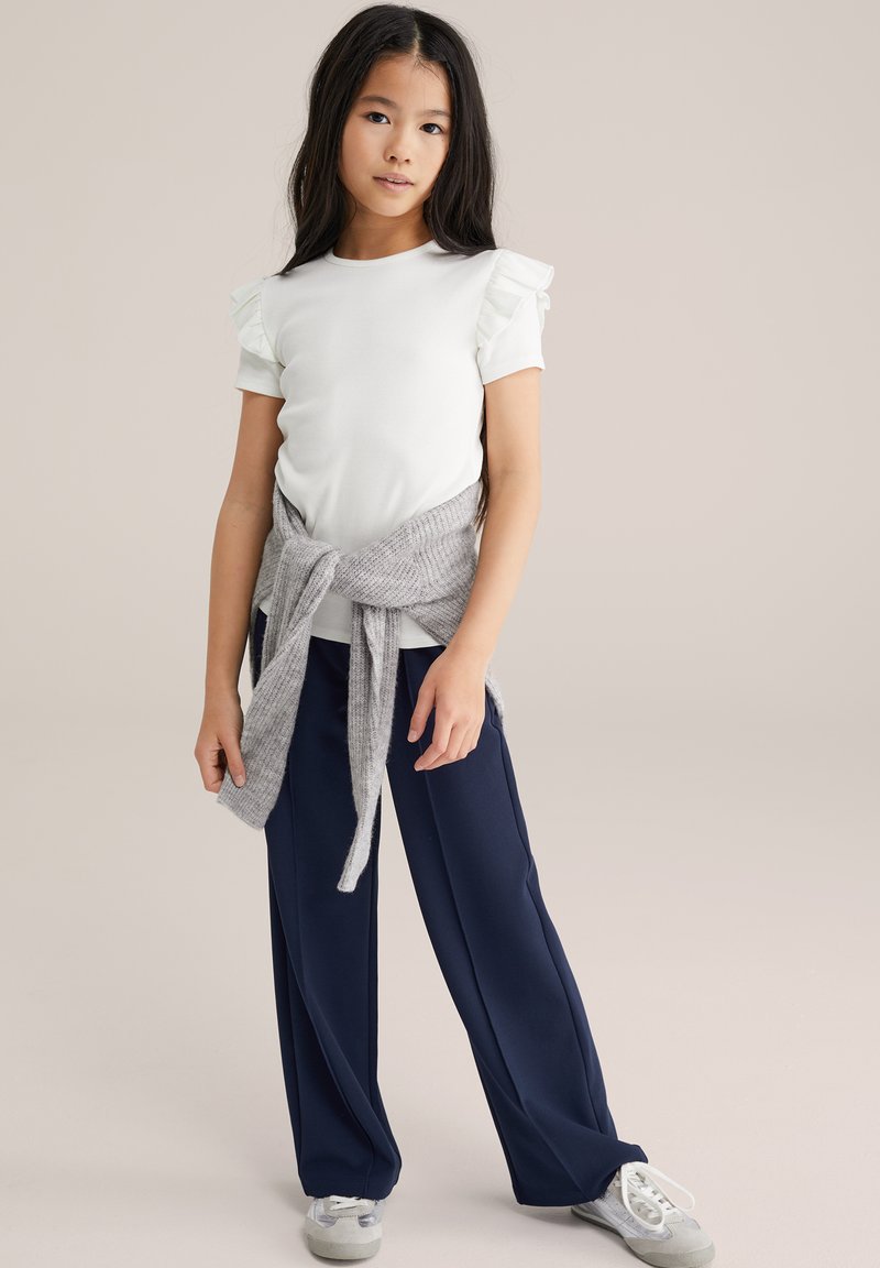 Mala djevojčica s dugom tamnom kosom koja nosi bijelu bluzu s volanima na rukavima, mornarsko plave široke hlače, sivi pulover vezan oko struka i bijele tenisice.