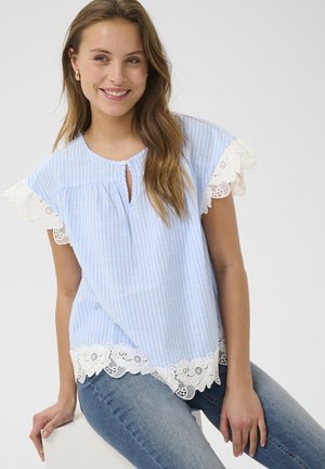 Ung kvinde smilende, iført en blå og hvidstribet bluse med hvide blonder på ærmer og kant, siddende på en hvid kube mod en ensfarvet baggrund.