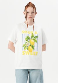 T-shirt bianca in cotone con una grafica colorata di limone e il testo "SOUR & SWEET" sotto. Vestibilità rilassata e maniche corte.