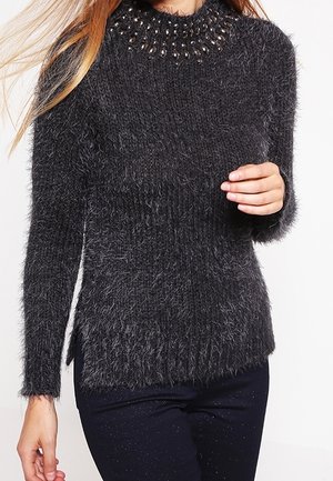 Femme portant un pull en maille grise foncée et duveteuse avec un col arrondi orné et des manches longues, associé à un pantalon foncé.