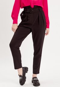 Schwarze hoch geschnittene Hose mit einem breiten Gürtel, Falten vorne und tapered Beinen, kombiniert mit schwarzen Slippern mit silberner Kettenakzent.