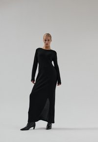 Czarna, prążkowana, dzianinowa sukienka maxi z długimi rękawami i bocznym rozcięciem, zestawiona z czarnymi, spiczastymi botkami, na prostym tle.