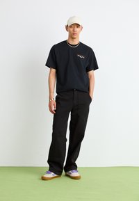 Black t-shirt with printed design, loose black pants, and cream cap. Footwear features purple accents and beige body with brown sole.