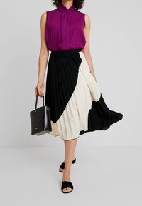 Schwarz-weißer plissierter Rock kombiniert mit einer lila ärmellosen Bluse. Eine schwarze Handtasche und schwarze Riemchensandalen runden das Outfit ab.