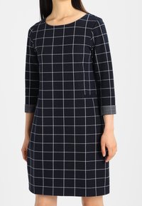 Robe bleu marine à motif quadrillé blanc, col rond, manches trois quarts avec poignets gris et poches latérales. Tissu en mélange de coton lisse.