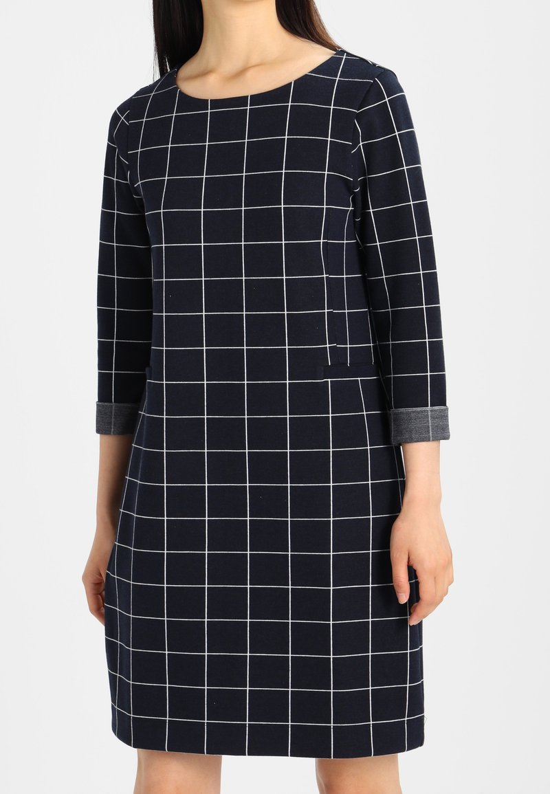 Robe bleu marine à motif quadrillé blanc, col rond, manches trois quarts avec poignets gris et poches latérales. Tissu en mélange de coton lisse.
