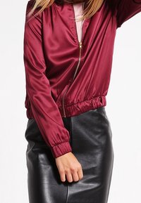 Burgundy bomber jacket made of shiny satin material, featuring a zip front, elastic cuffs, and a gathered hem, paired with a black leather skirt.