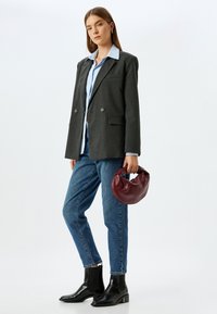 Blazer gris de corte cruzado, camisa azul claro por debajo, jeans azules, botas negras hasta el tobillo, sosteniendo un bolso circular burdeos.