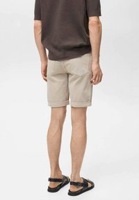 Beige Shorts mit umgeschlagenen Säumen, ausgestattet mit Gesäßtaschen und dezenten Stickdetails, kombiniert mit schwarzen Sandalen. Lässiger, leichter Stoff.