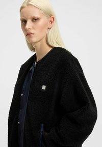 Black textured bomber jacket with a plush fabric, featuring a navy inner lining and silver snap buttons, alongside a small branded badge.