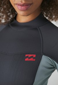 En våtdräkt med en åtsittande design, svarta och turkosa paneler, rundad halsringning, flatlock-sömmar och en röd logotypaccent på bröstet.