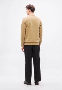 Béžová mikina s okrúhlym výstrihom a rebrovanými manžetami, nosená s čiernymi širokými nohavicami a topánkami. Pohľad zozadu, hladká textúra, minimalistický dizajn.