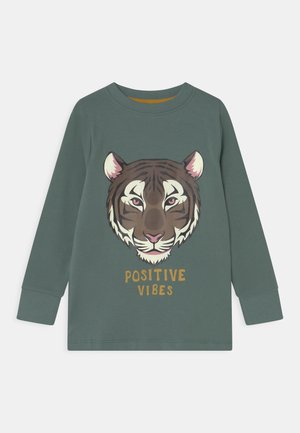 Groen shirt met lange mouwen met een tijgergezicht en de woorden "POSITIVE VIBES" gedrukt onder de illustratie.