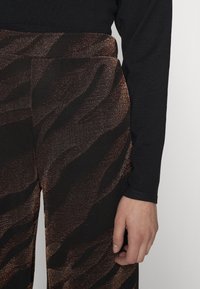 Pantalons noirs avec un motif rayé zebra en bronze scintillant, offrant une texture lisse et un design à jambes larges. Main partiellement visible.