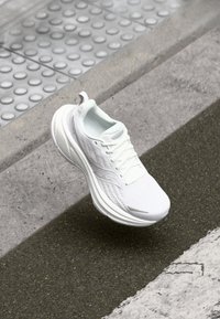 Hvid atletisk sko med et mesh-oberste, struktureret design og en tyk sål. Har en glat tåboks og snøresystem.