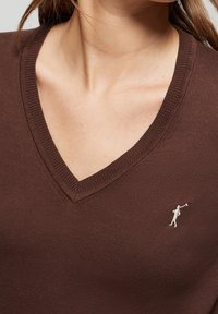 Pull marron à col en V avec encolure côtelée ; présente un petit logo argenté sur le côté gauche ; fabriqué en tissu tricoté lisse.