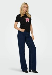 Lys hår kvinde iført sort grafisk t-shirt med pink sløjfe-design, mørkeblå brede bukser og sorte ankelstøvler, står med den ene hånd i lommen.