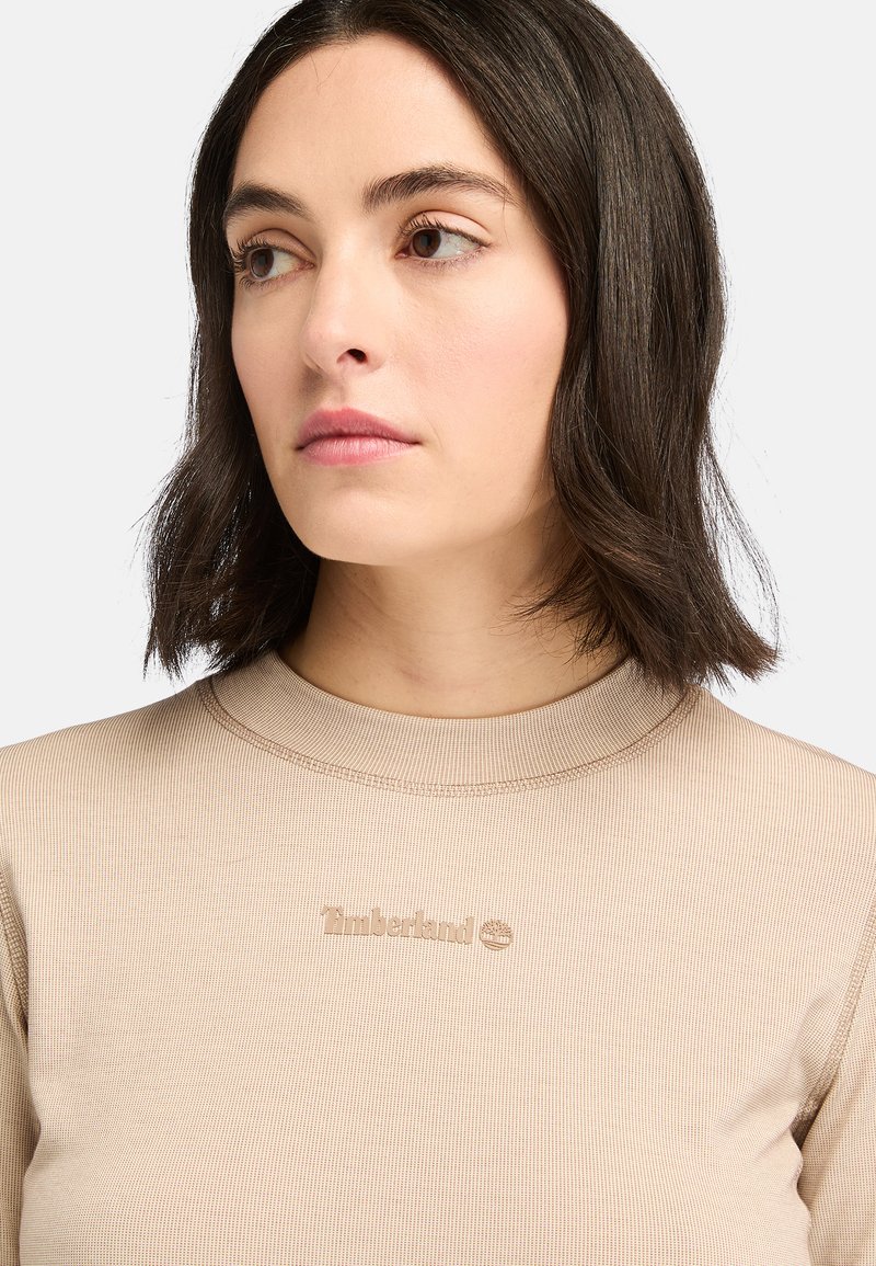 Top beige a maniche lunghe con righe, caratterizzato da un collo alto e un sottile logo Timberland tono su tono sul petto. Texture liscia.