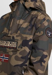 Camo-gepatrooneerde jas in tinten groen en bruin, met een rits aan de voorkant, een groot borstzak met branding en een logo-patch op de mouw.