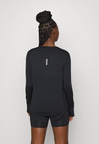 Kobieta z warkoczami związanymi w kok, nosi czarną, długą koszulkę sportową i czarne szorty, widziana od tyłu na jednolitym tle.