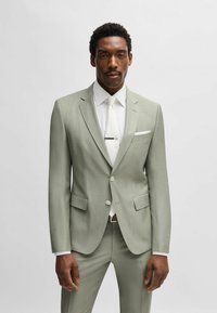 Abito verde chiaro con design su misura, con revers a forma di notti, due tasche frontali e una camicia bianca con cravatta e fermacravatta.