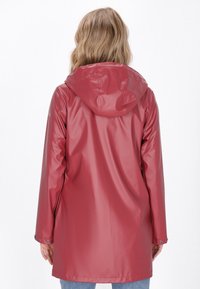 Punainen vedenpitävä sadetakki hupulla, pitkät hihat ja nappikiinnitys. Sileä, kiiltävä pinta; polviin ulottuva malli.