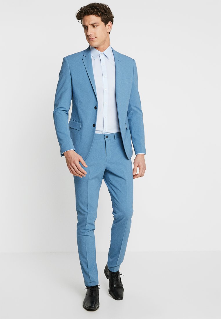 Traje azul claro que incluye una chaqueta entallada con un solo botón, pantalones a juego y una camisa blanca de botones. Completado con zapatos negros.