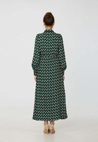 Robe longue verte avec un motif géométrique rose et blanc. Elle présente des manches longues, une taille cintrée et une jupe fluide.