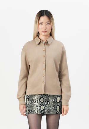 Beige skjorte med knappelukking og krage, som har syv knapper, i kombinasjon med et strukturert svart-hvitt slangeskinnsmønster skjørt. Glatt stoff.