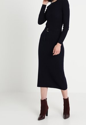 Kobieta ubrana w czarną, prążkowaną sukienkę midi na długi rękaw z paskiem, zestawioną z ciemnobrązowymi, wysokimi botkami na obcasie na jednolitym tle.