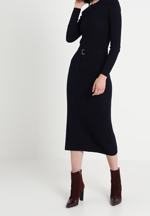 Femme portant une robe midi noire à manches longues côtelée avec une ceinture, associée à des bottines à talons hauts marron foncé sur un fond uni.