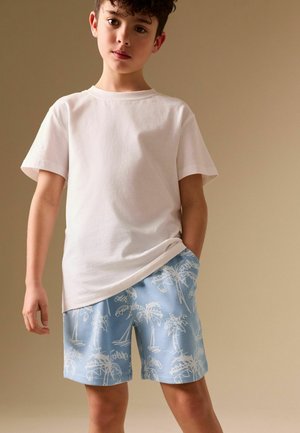 Boy with curly hair wears plain white t-shirt and light blue shorts with white palm tree print, standing against beige background.