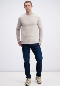 Beżowa bluza z długim rękawem z prążkowanym dekoltem i mankietami, zestawiona z ciemnoniebieskimi dżinsami i beżowymi trampkami z unikalnym wzorem.