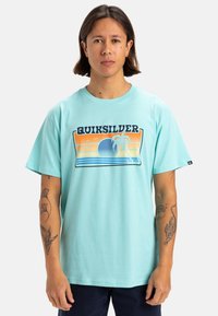 Camiseta de algodón turquesa con un estampado gráfico que presenta un diseño de atardecer y palmera. Manga corta y estilo de cuello redondo.