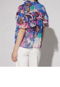 Blouse légère et transparente avec des manches volantes, ornée d'un motif floral multicolore dans les tons rose, bleu et marron. Associée à un pantalon blanc.