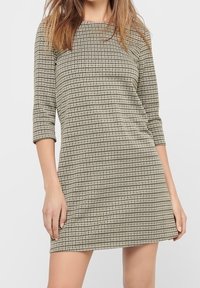 Kontrollerad klänning i beige och svart, med ärmar som når till armbågen och en rak silhuett. Texturerad tyg med ett subtilt mönster.