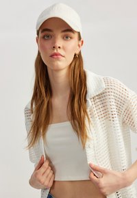 Cárdigan blanco de punto abierto superpuesto a un top corto de canalé blanco, combinado con una gorra de béisbol blanca. Cabello largo, pendientes de aro dorados sutiles visibles.