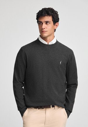 Pull en tricot gris foncé avec un col rond et des manches longues, présentant un petit logo blanc sur la poitrine gauche. Texture lisse.