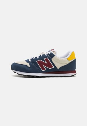 Scarpa da ginnastica blu navy e beige con lacci bianchi, logo "N" color vinaccia, dettaglio giallo sul tallone e suola in gomma nera su sfondo bianco.