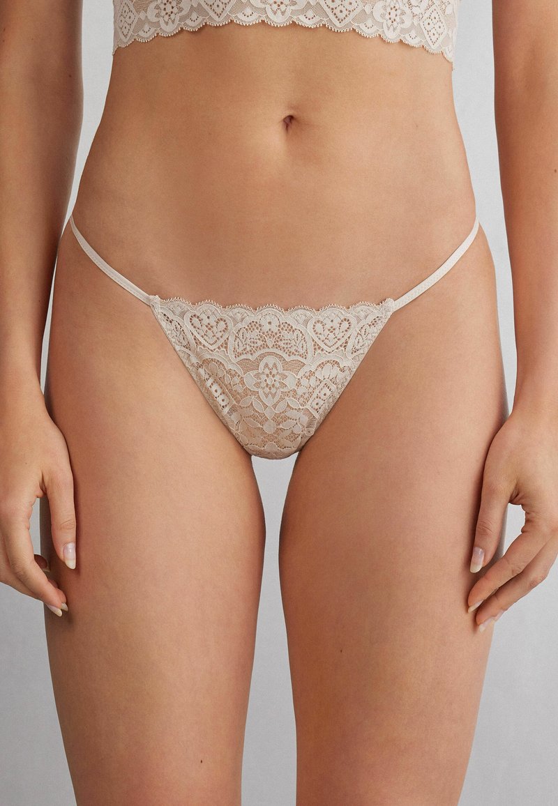 String en dentelle beige clair, présentant des bords festonnés, des motifs floraux délicats et des fines lanières sur les côtés pour une couverture minimale.