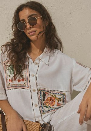 Hosszú, göndör hajú nő, kerek napszemüveget visel, fehér, gombos ingben, színes hímzett mintákkal, kezében egy fonott táskával.