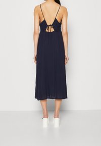 Námorně modré midi šaty s tenkými ramínky, otevřeným zády svázanými v pase, splývavou látkou a mírně průsvitnou texturou. V kombinaci s bílými botami na blokovém podpatku.