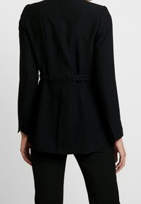 Blazer negro a medida con cintura entallada, hombros estructurados, mangas largas y pliegues en la parte trasera. Tejido suave con costuras mínimas visibles.