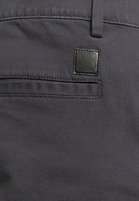 Grå tygbyxor med en slät textur, med en rektangulär svart läderlapp med ett präglat "B"-logotyp och en bakfickedesign.