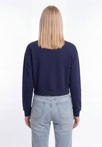 Egyenes szőke hajú nő sötétkék hosszú ujjú, rövid felsőt visel és világoskék, magasderekú farmert, háttal a nézőnek.