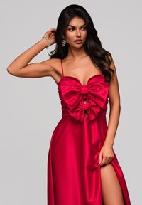 Abito in satin rosso caratterizzato da un busto aderente con grandi fiocchi e dettagli cut-out, gonna fluida con uno spacco alto fino alla coscia, spalline sottili.