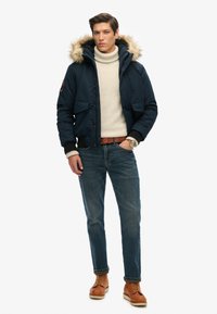 Marineblå puffjakke med en pelsforet hætte, kombineret med en cremefarvet turtleneck sweater, blå jeans og brune snørebåndsstøvler. Bemærkelsesværdige lommer på jakken.