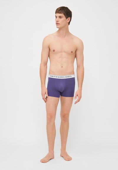 Ljubičaste pamučne bokserice s bijelim pojasom na kojem stoji logo "POLO RALPH LAUREN". Nosene od strane modela koji stoji bosi na bijeloj pozadini.