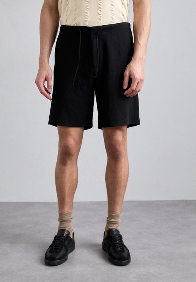 Schwarze Baumwollshorts mit elastischem Bund und Zugband. Glatte Textur, knielanger Schnitt, kombiniert mit schwarzen Sneakers und beigen Socken.