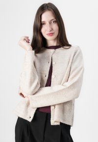 Licht beige cardigan van zacht, textuurmateriaal. Heeft een ronde halslijn, vijf knopen en lange mouwen. Draag het over een donkere top.